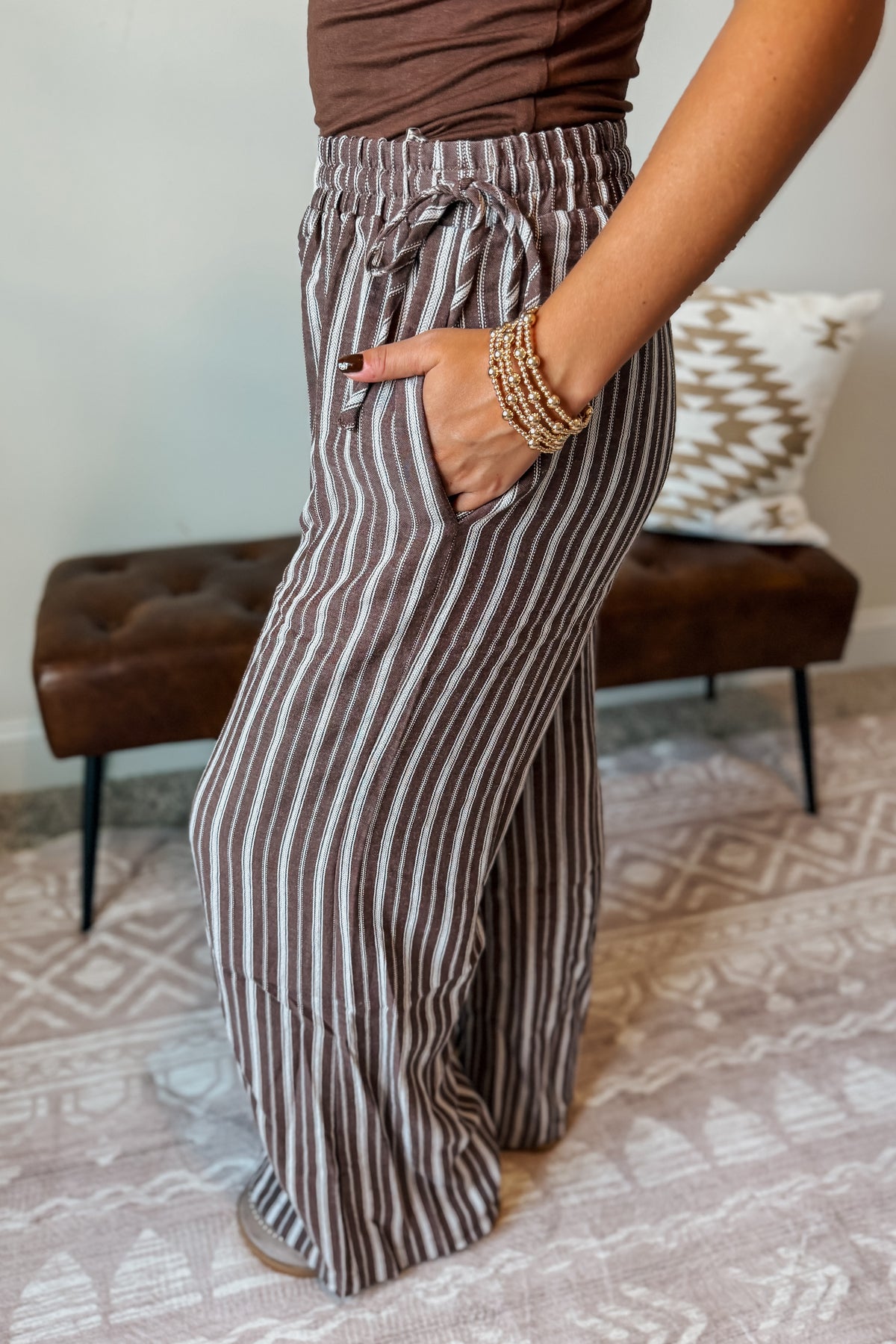 A person wearing brown and beige vertically striped wide-leg pants with a drawstring waist, standing in a living room setting with a patterned rug and a brown leather bench in the background.