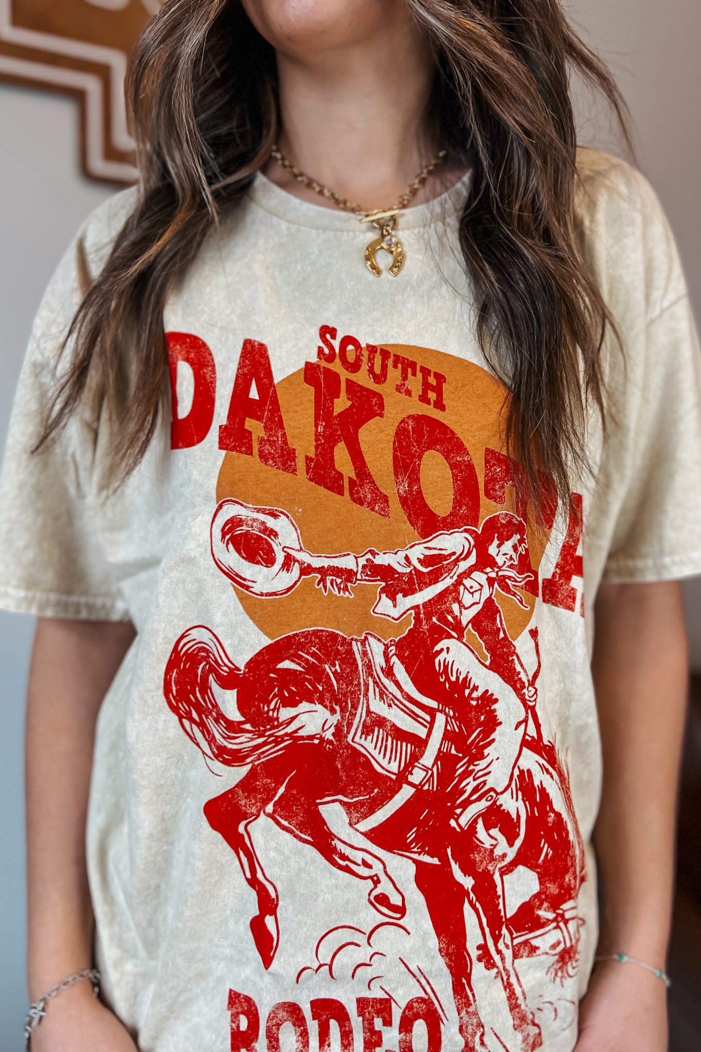Light tan mineral-wash T-shirt with red “South Dakota Rodeo” graphic featuring a cowboy riding a bucking horse inside an orange sunset design