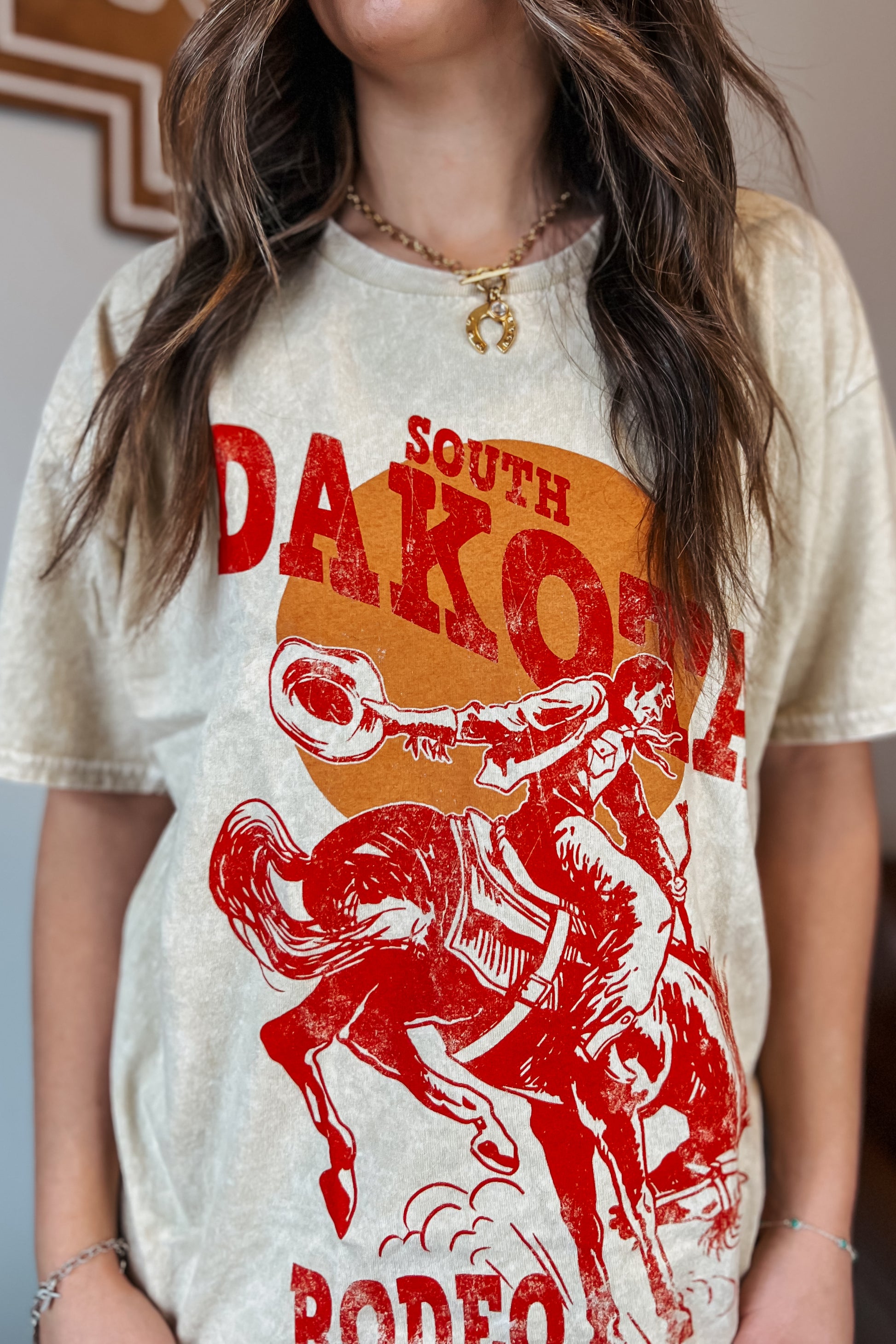 Light tan mineral-wash T-shirt with red “South Dakota Rodeo” graphic featuring a cowboy riding a bucking horse inside an orange sunset design