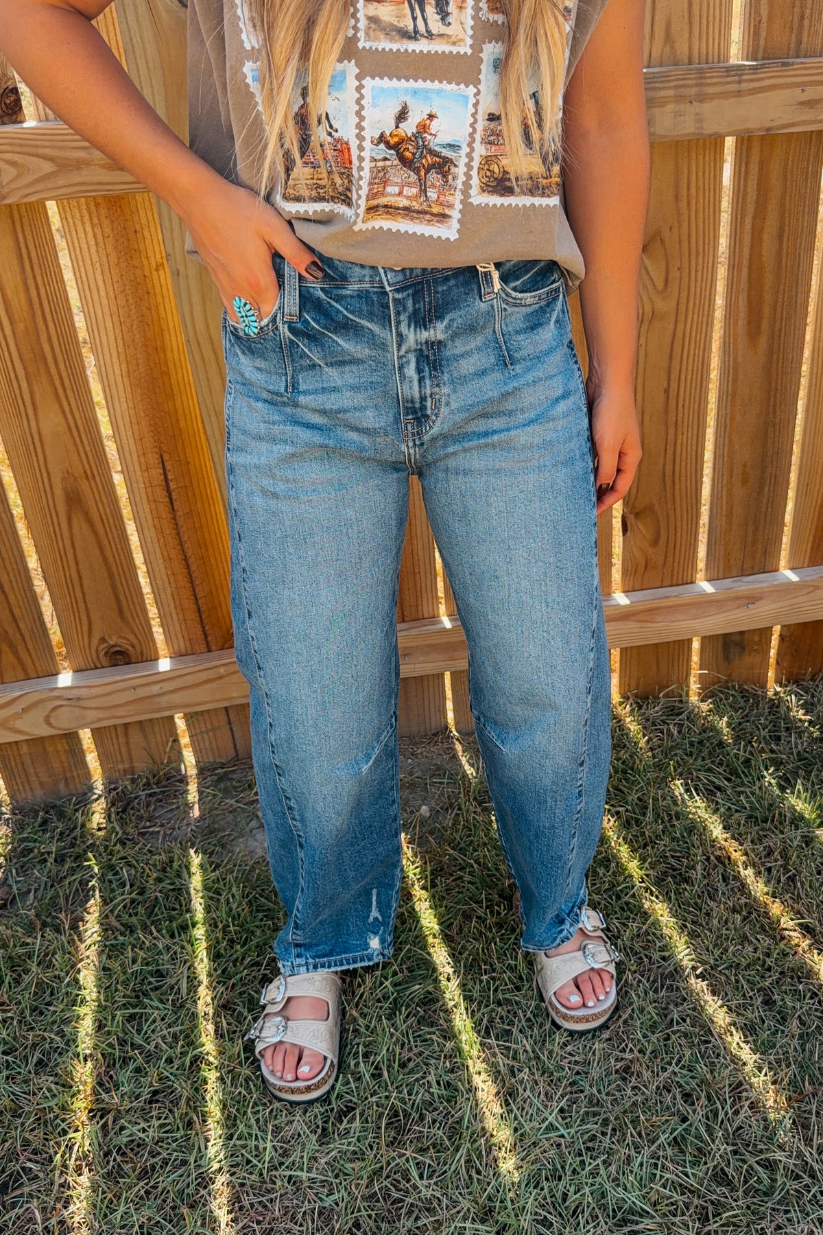 A person standing outdoors in front of a wooden fence and grass, wearing dark-wash blue jeans, a graphic t-shirt, and sandals.