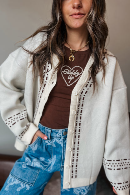 Model wearing a cream knit cardigan with brown western-inspired stitch detailing over a brown graphic tee and blue western print jeans.