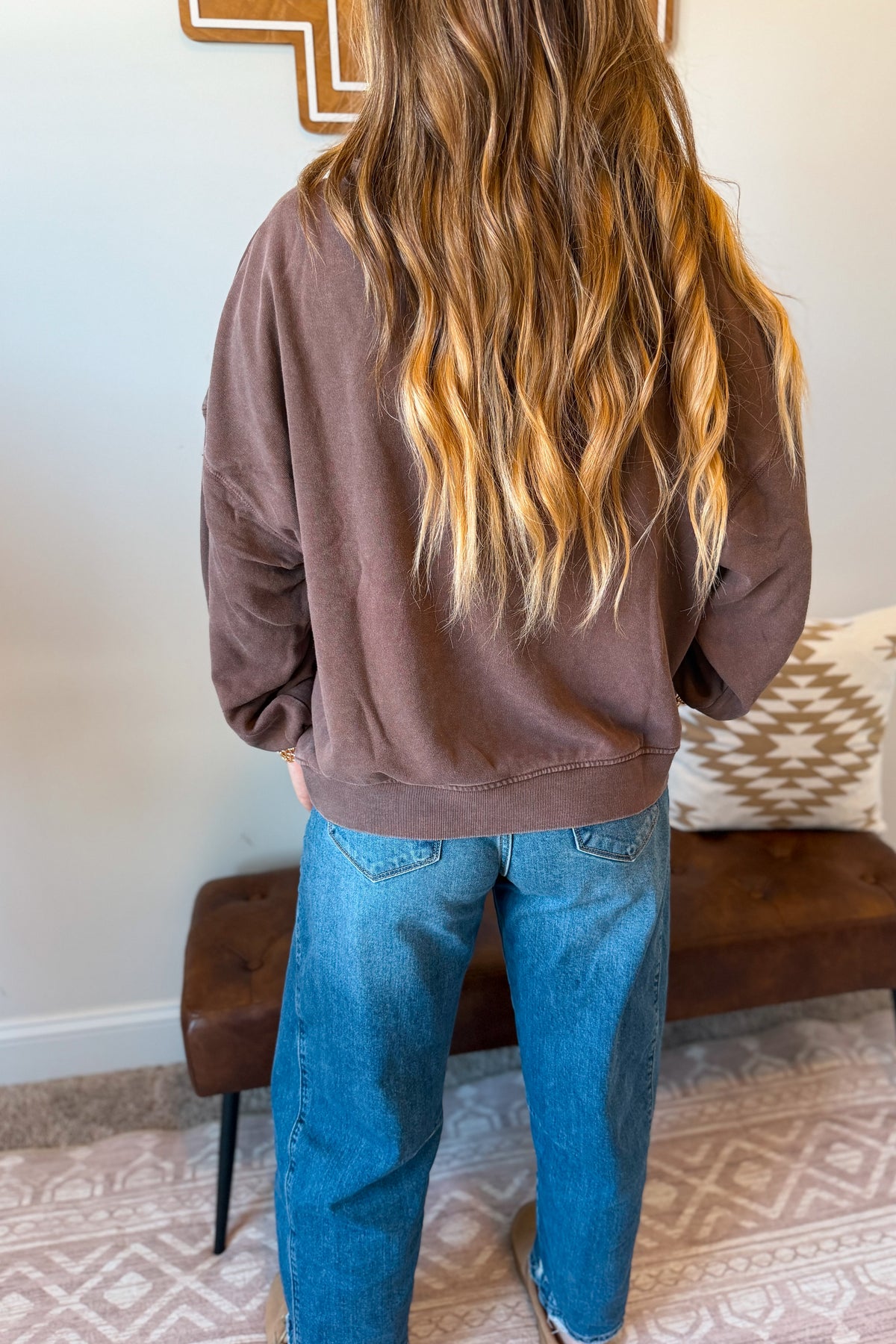 A person viewed from behind wearing a brown long-sleeve sweatshirt and blue jeans, standing in a home interior with a bench, patterned rug, and wall decor visible.