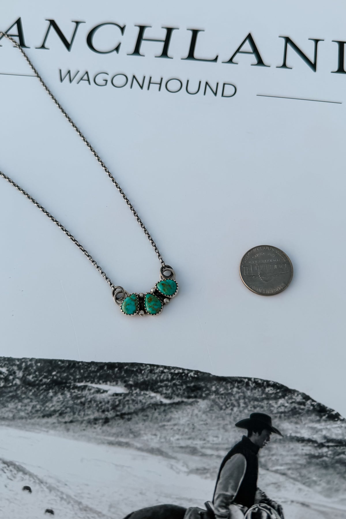A close-up of a necklace with three small turquoise-like green stones in a silver-toned setting on a chain, placed on top of a white book