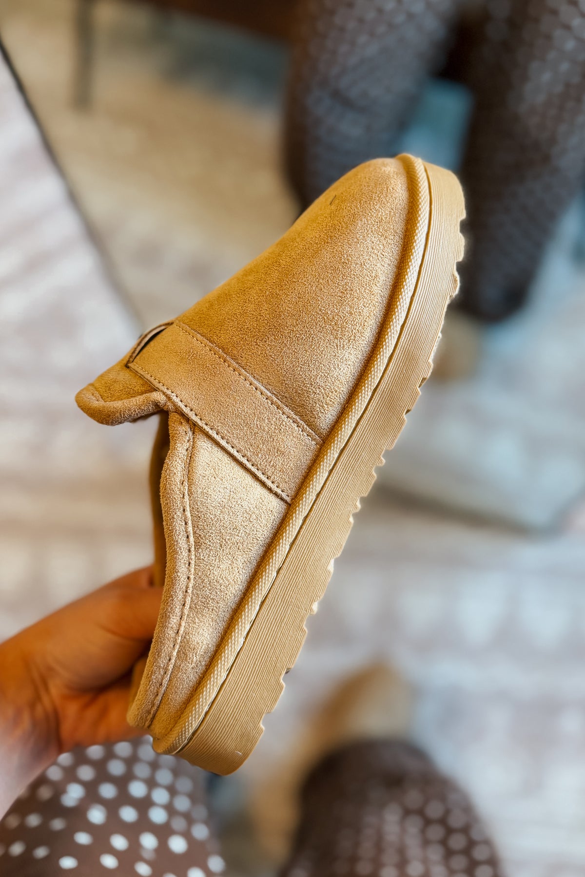 A tan faux-suede slip-on shoe held in a hand, showing the side profile with a thick lug sole, against a blurred indoor background.