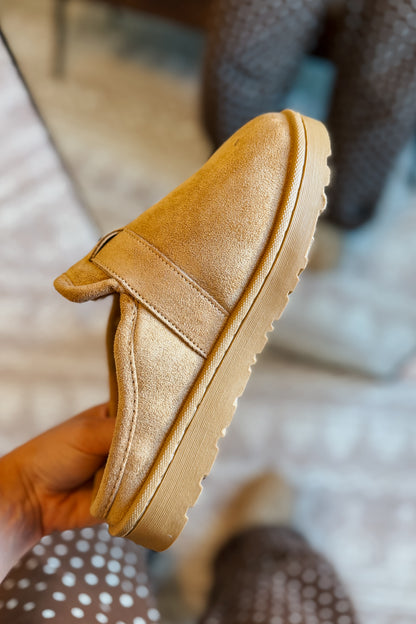 A tan faux-suede slip-on shoe held in a hand, showing the side profile with a thick lug sole, against a blurred indoor background.