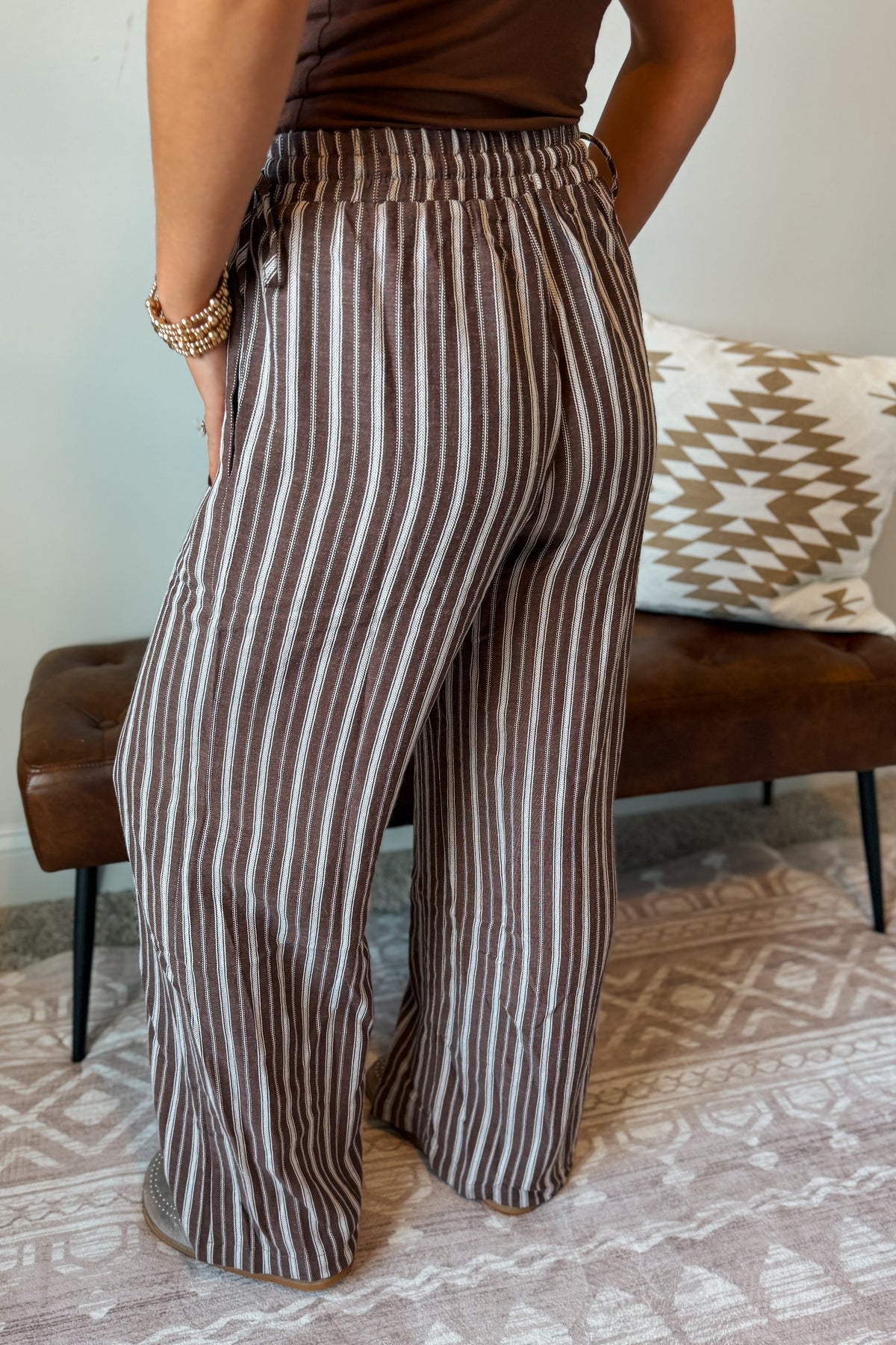 A person wearing high-waisted, wide-leg pants with vertical brown and beige stripes, standing in a living room setting with a patterned rug and a brown leather bench in the background.