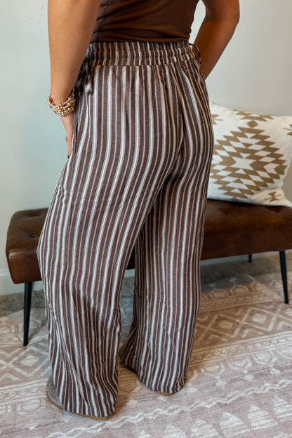 A person wearing high-waisted, wide-leg pants with vertical brown and beige stripes, standing in a living room setting with a patterned rug and a brown leather bench in the background.