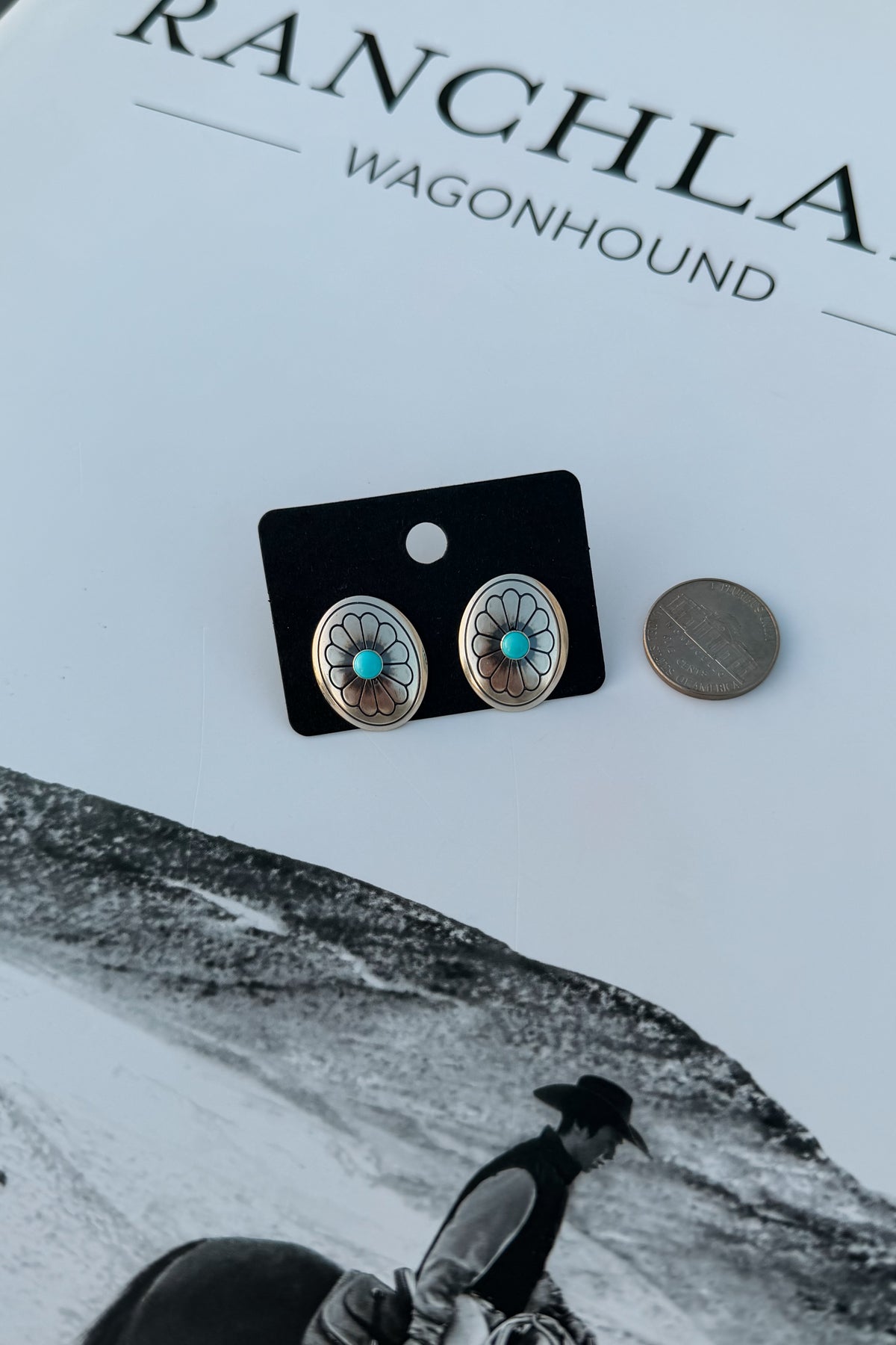 A pair of oval stud earrings with turquoise centers and silver-toned floral detailing, displayed on a black card next to a coin for scale