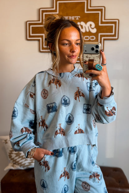 A woman takes a mirror selfie wearing a light blue matching lounge set with a brown and blue cowboy and rider print. The set includes a cropped hoodie and sweatpants, shown in a home interior setting.