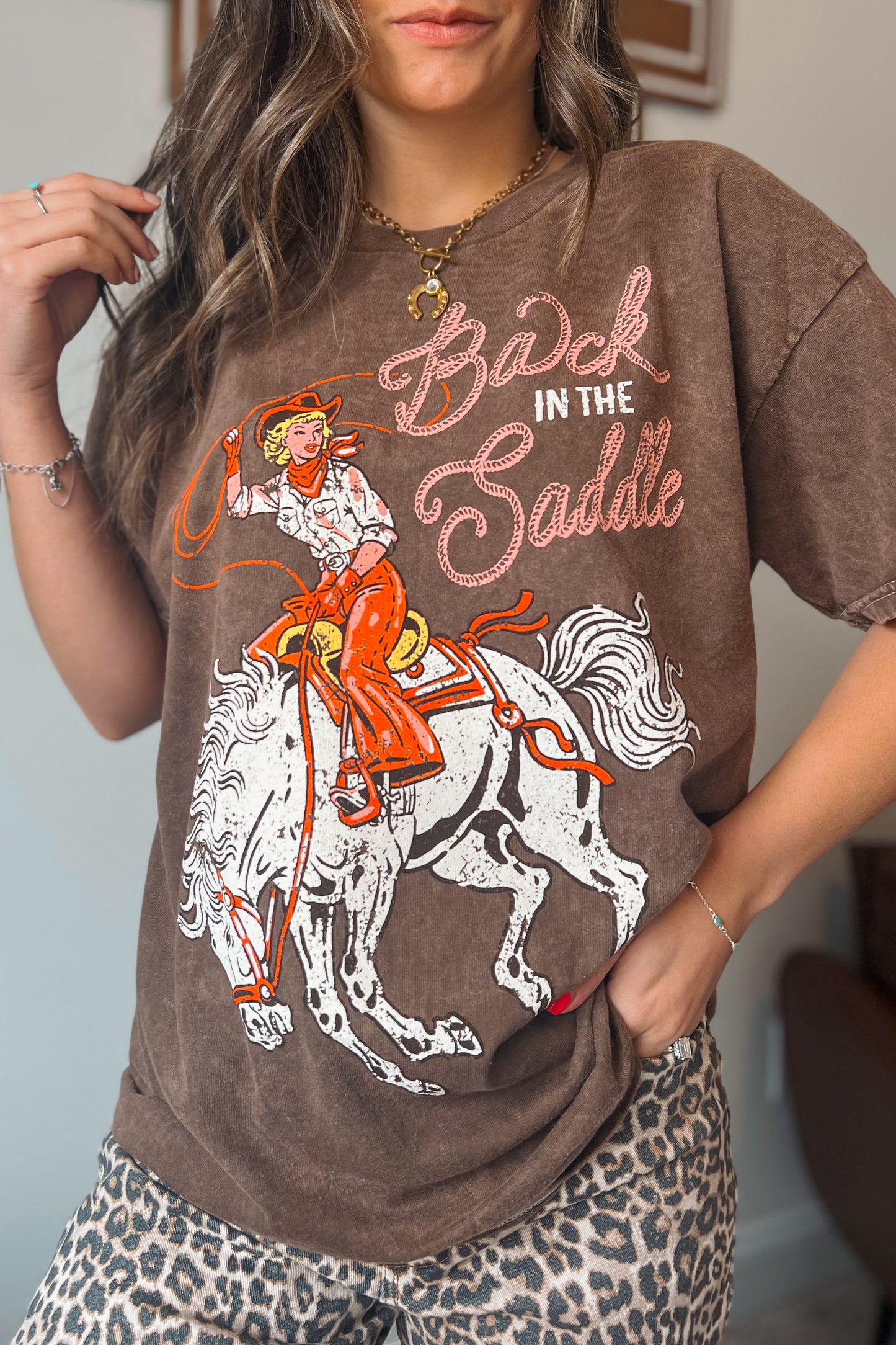 Brown vintage-wash graphic tee featuring a cowgirl riding a horse with “Back in the Saddle” text, styled with leopard print bottoms.