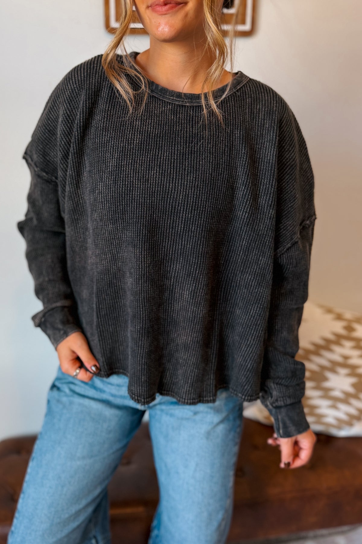 A woman wearing a dark charcoal gray ribbed knit long-sleeve top and light blue jeans, standing indoors.