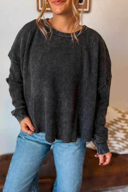 A woman wearing a dark charcoal gray ribbed knit long-sleeve top and light blue jeans, standing indoors.