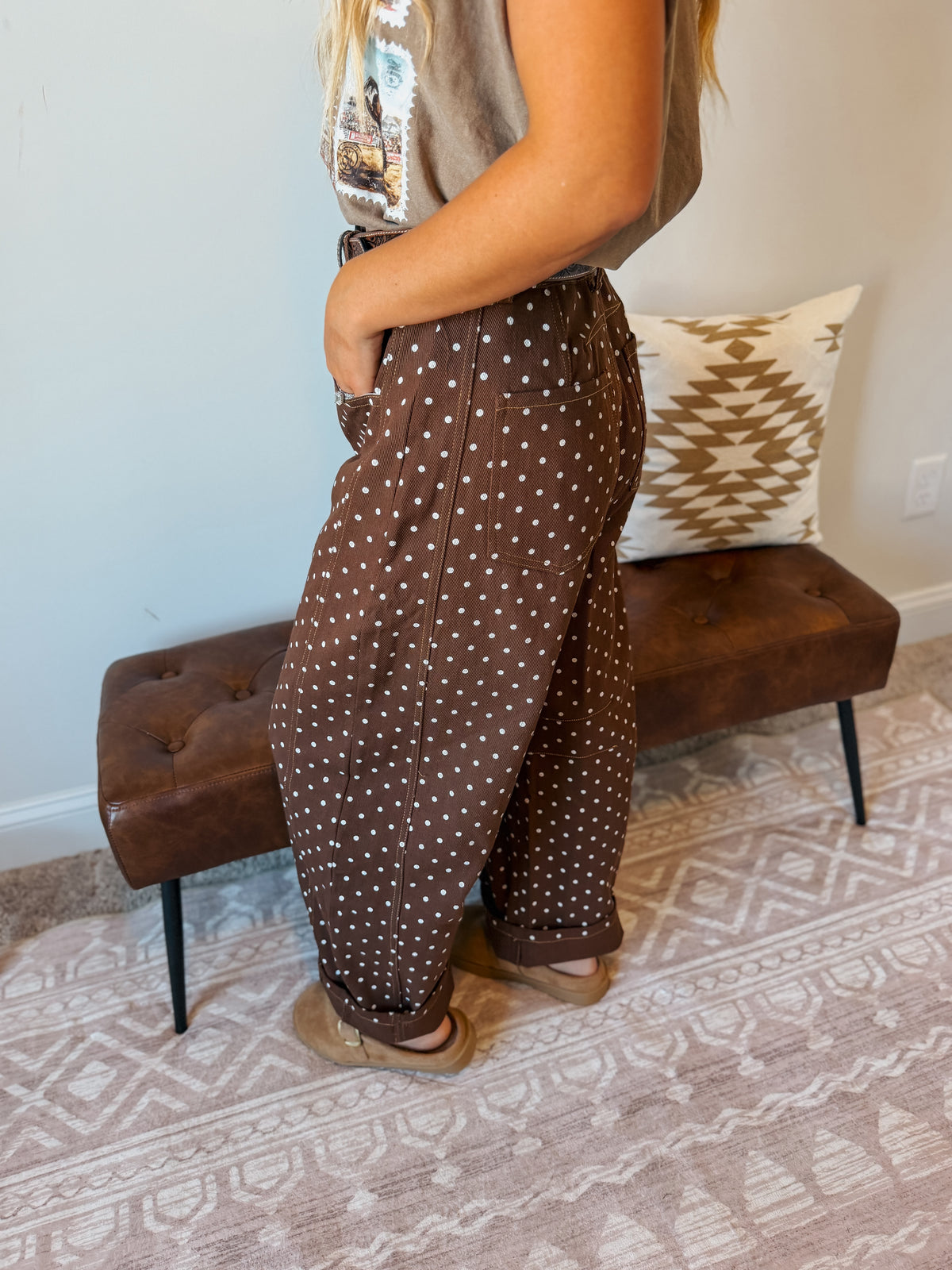 A person wearing brown wide-leg pants with white polka dots, standing in a room with a tufted brown bench and patterned rug.