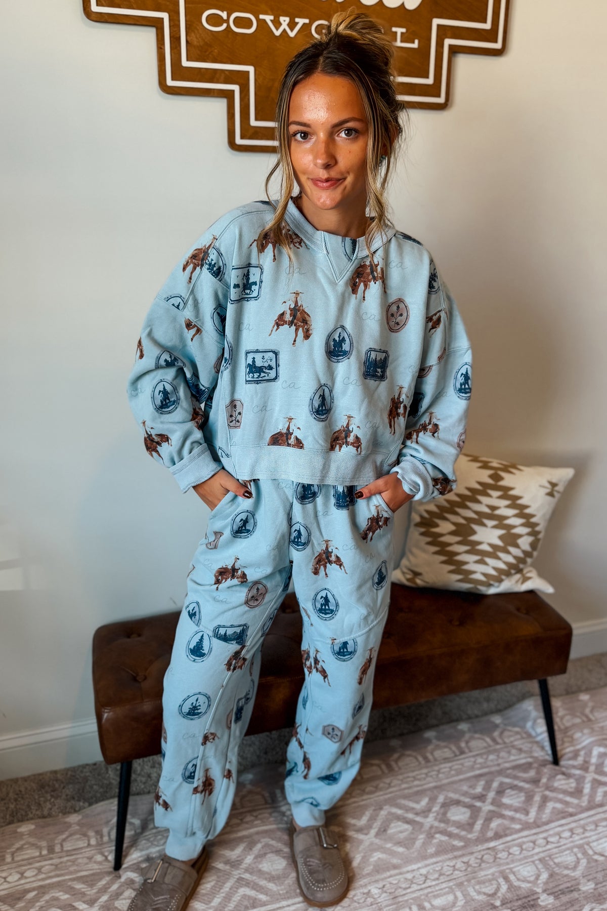 A woman stands indoors wearing a matching light blue lounge set with a brown and navy lobster print. The set includes a long-sleeve top and jogger pants, styled with brown slip-on shoes.