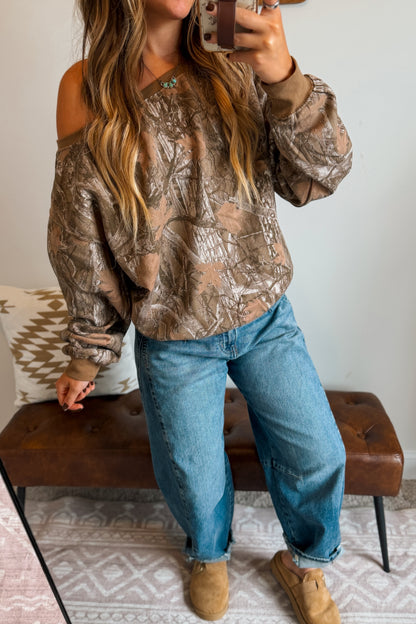 A woman takes a mirror selfie wearing a brown and beige camouflage-patterned off-the-shoulder sweatshirt, blue jeans, and tan suede boots, sitting on a brown leather bench.
