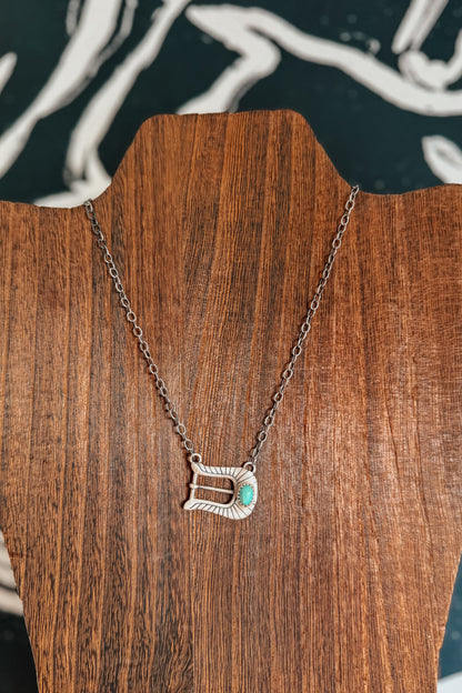 A silver-toned pendant necklace with a textured, openwork design and a small turquoise accent, displayed on a brown wooden stand.
