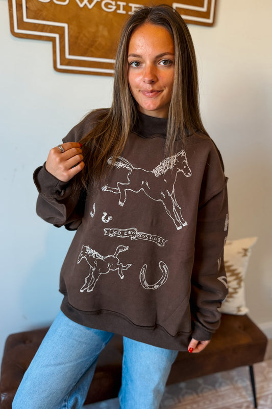 A woman wearing a brown crewneck sweatshirt with white western-style horse illustrations and blue jeans, posing in an indoor setting.