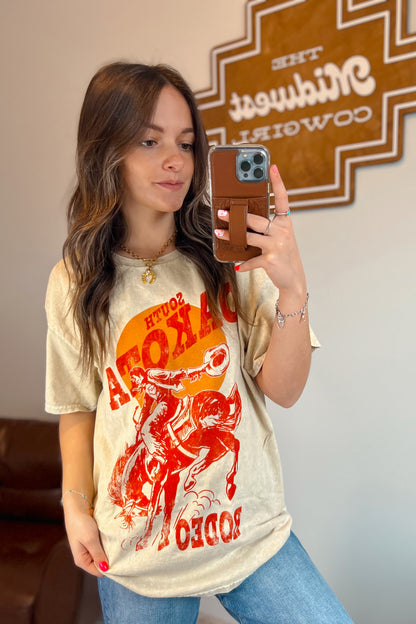 Light tan mineral-wash T-shirt with red “South Dakota Rodeo” graphic featuring a cowboy riding a bucking horse inside an orange sunset design