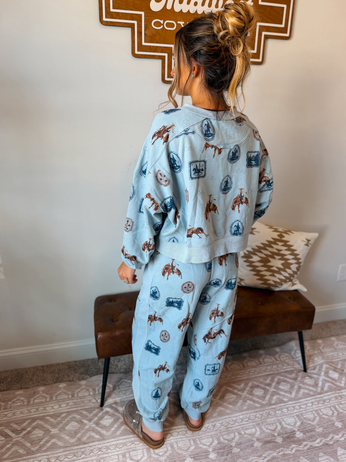 A woman stands with her back to the camera wearing a light blue matching set with a brown and blue western-themed print, consisting of a long-sleeve top and jogger pants.