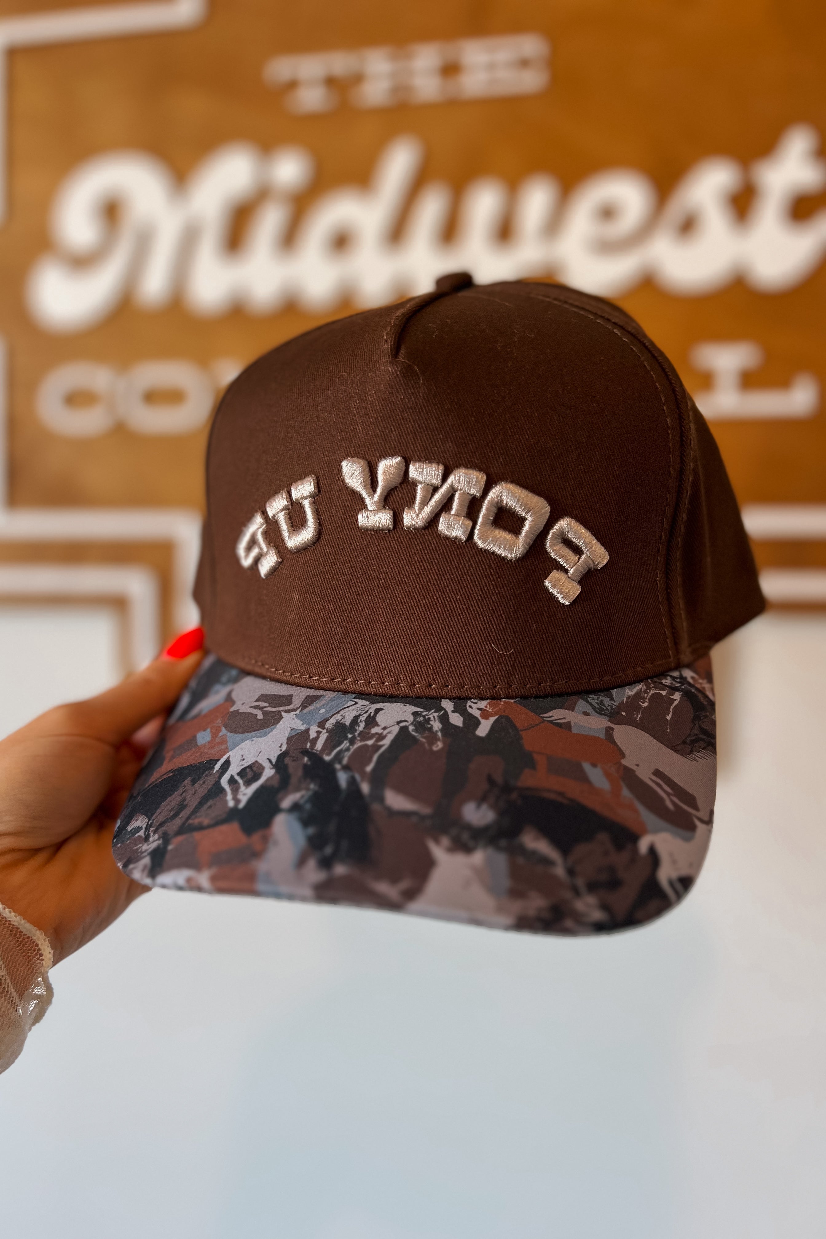 Brown trucker hat with embroidered “Pony Up” text in cream lettering and color block horse print on the bill.