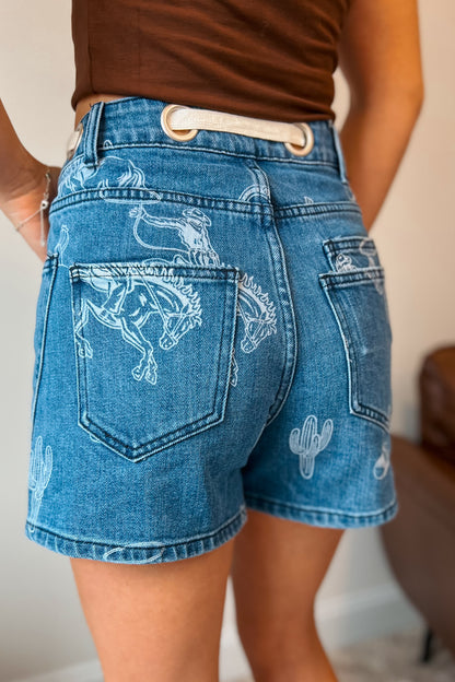 Medium wash denim shorts with bleached western print featuring bucking horses, cowboys, cacti, and horseshoes. High-rise fit with front and back patch pockets, belt loops, and cream rope drawstring threaded through large metal grommets at the waistband.