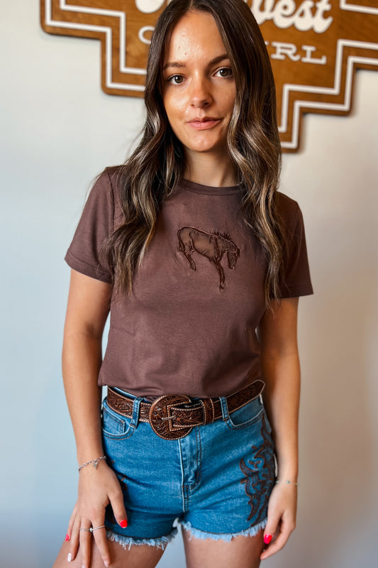Brown fitted graphic tee with embroidered bucking horse design, styled with a brown tooled leather belt and light wash denim shorts with western embroidery.