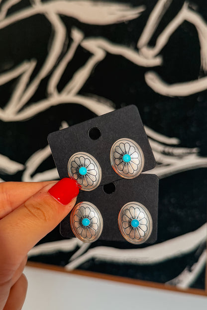 A hand with red nail polish holds a black display card featuring four silver-toned oval stud earrings with turquoise centers and floral inlay details, against a dark patterned background.