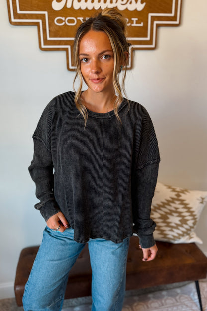 A woman wearing a dark charcoal gray ribbed long-sleeve crewneck sweatshirt and light blue jeans, standing in a home setting with a patterned pillow and a wooden sign in the background.
