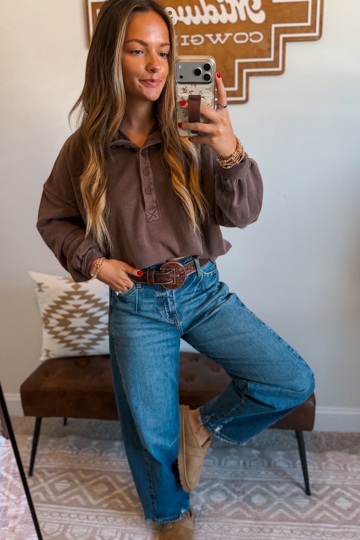 A woman takes a mirror selfie wearing a brown long-sleeve henley shirt and blue jeans with a brown belt, standing in a room with a patterned rug and a bench.