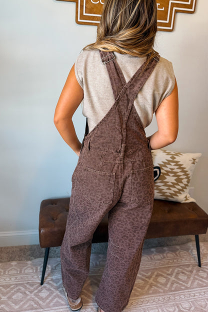 A woman viewed from behind wearing brown leopard-print overall jumpsuit over a light beige sleeveless top, standing in a home setting with a brown leather bench and patterned rug.