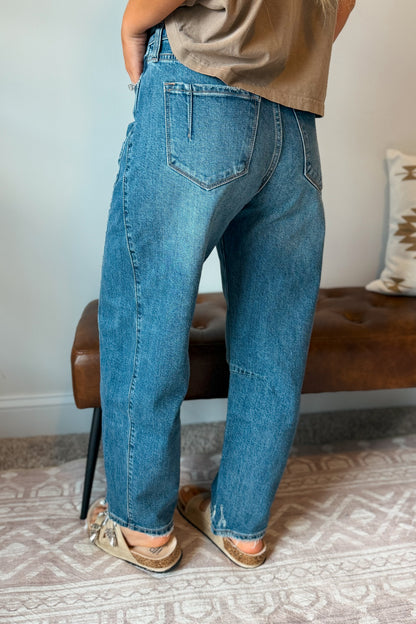 A person shown from the back wearing loose-fit medium-wash blue jeans, a tan top, and tan sandals, standing indoors near a brown leather bench and patterned rug.