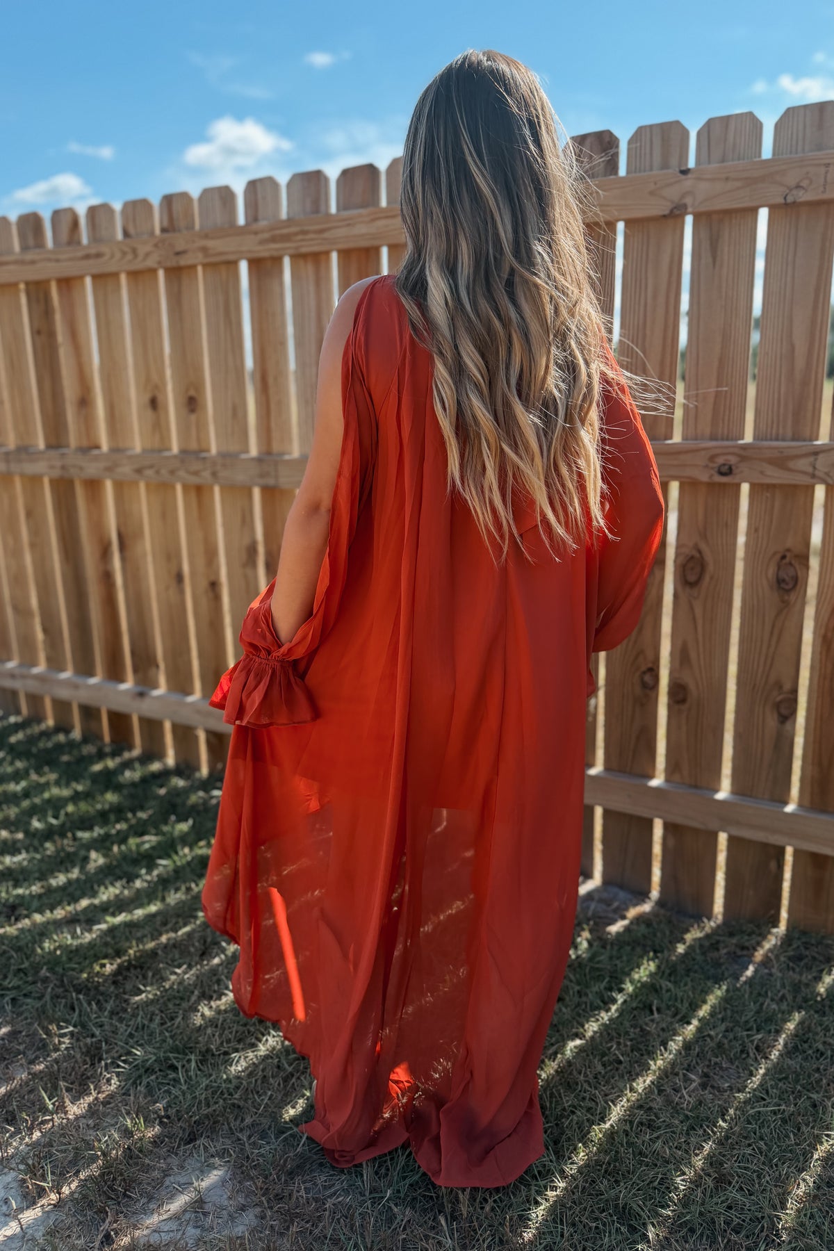 A woman viewed from behind wearing a flowing rust-orange sheer maxi dress with tie details at the sleeves, standing outdoors in front of a wooden fence under a blue sky.