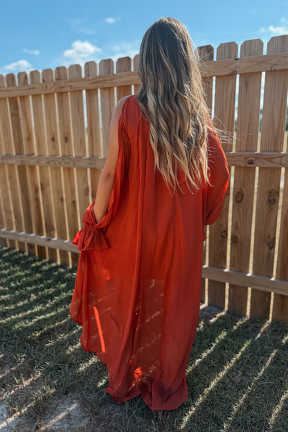 A woman viewed from behind wearing a flowing rust-orange sheer maxi dress with tie details at the sleeves, standing outdoors in front of a wooden fence under a blue sky.