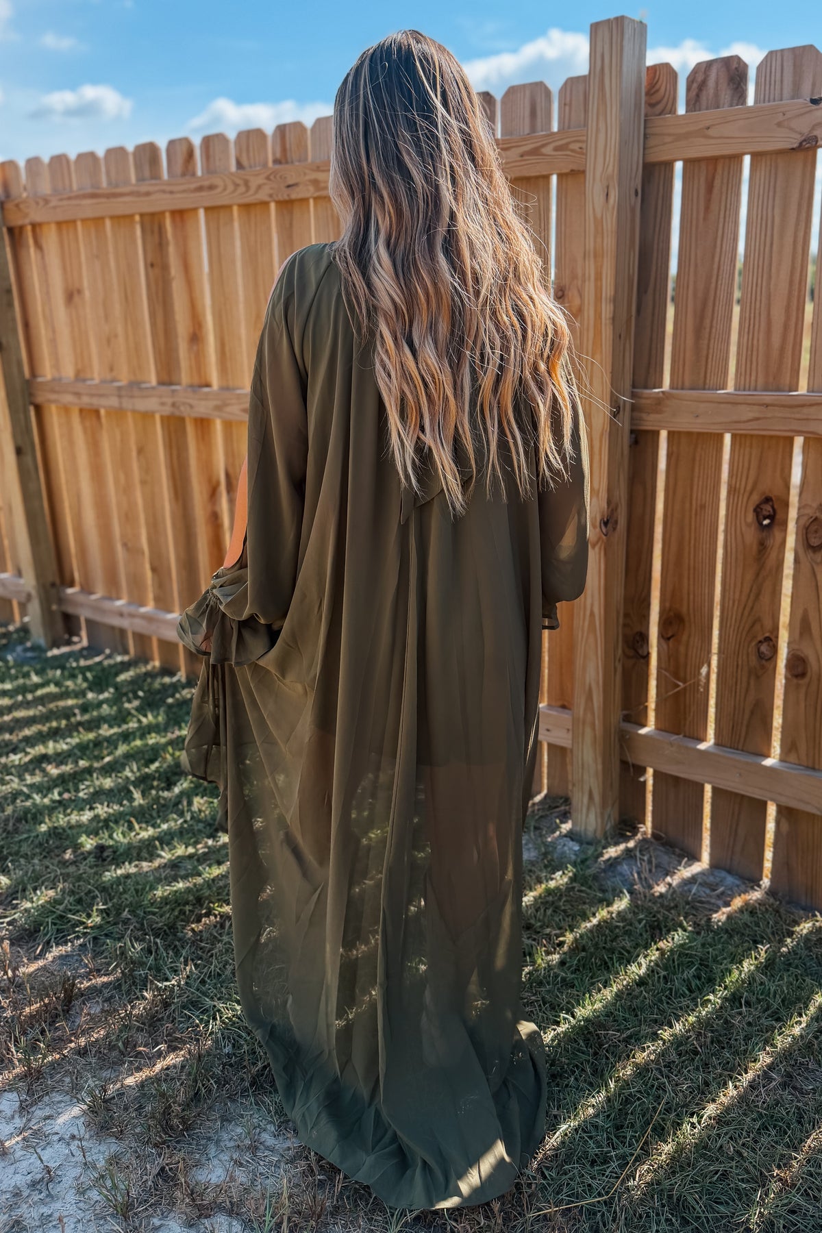 A woman viewed from behind wearing a long, flowing olive green kaftan dress in an outdoor backyard setting with a wooden fence and grass.