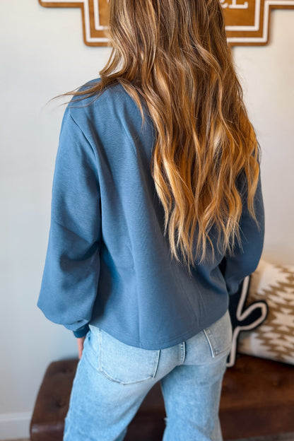 A woman viewed from behind wearing a dusty slate blue long-sleeve top and light wash blue jeans in a home setting.
