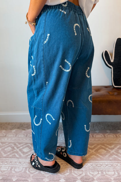 A person wearing loose blue denim pants with a white horseshoe pattern, standing indoors on a patterned rug and wearing black slide sandals.