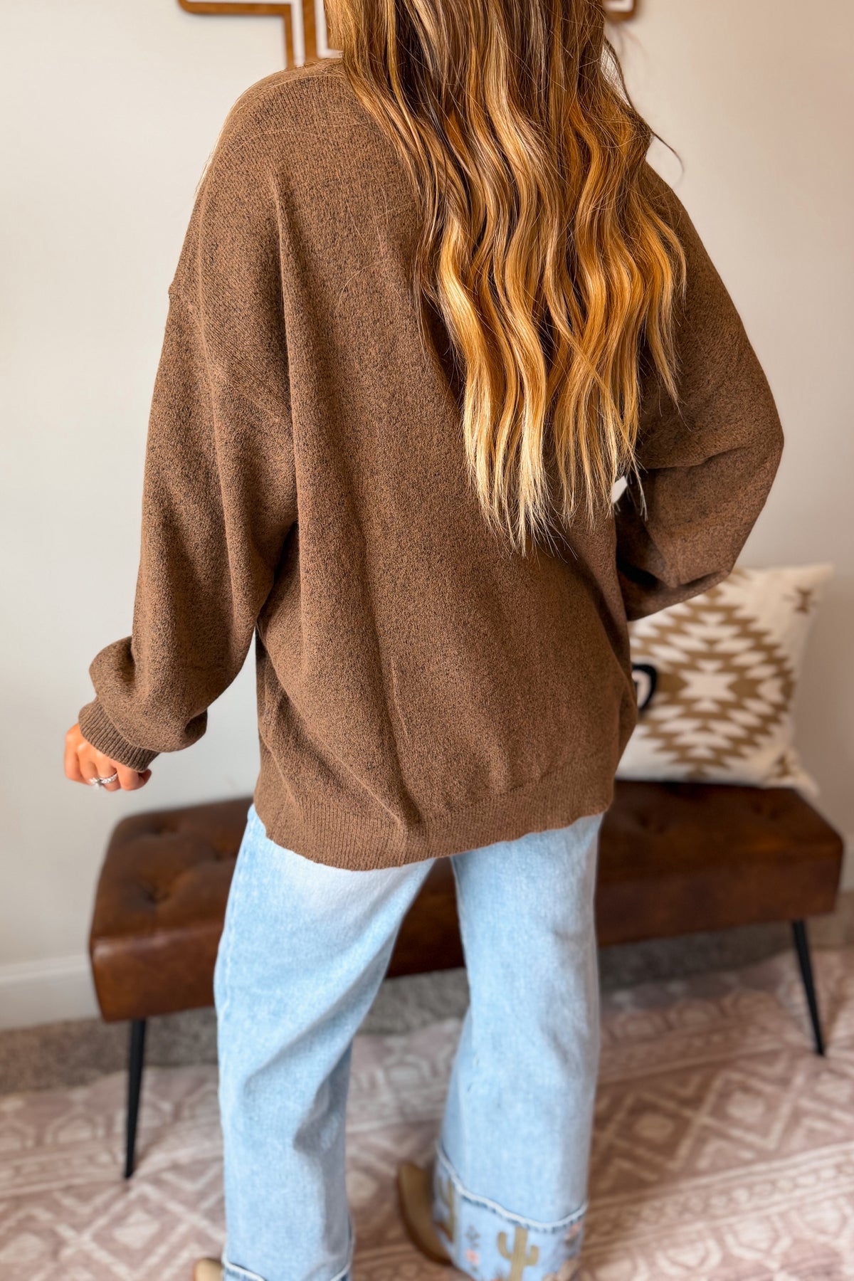 A woman shown from behind wearing a brown knit sweater and light blue jeans in a home setting with a tufted bench and patterned rug.