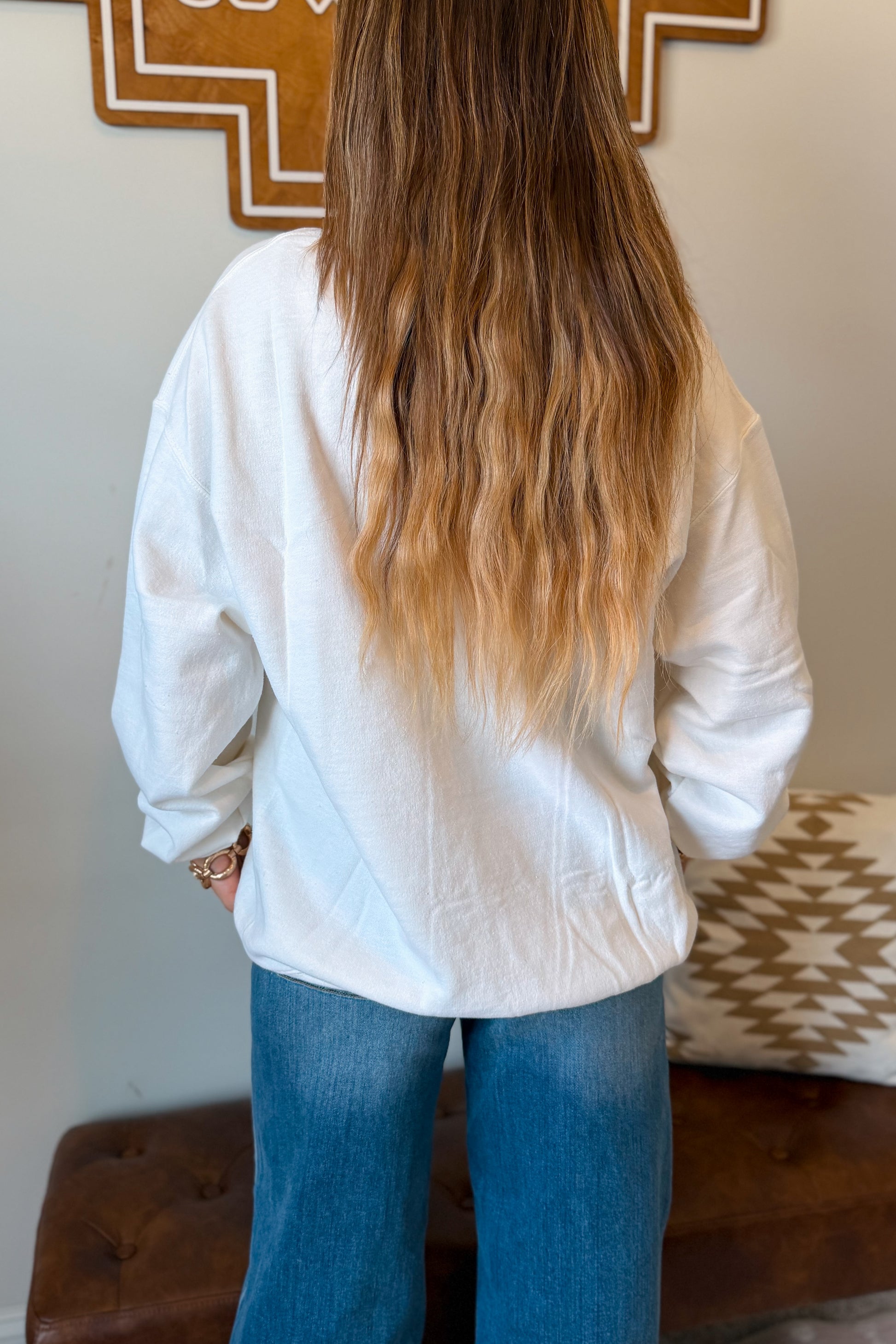 A woman viewed from behind wearing a loose white long-sleeve shirt and blue jeans, standing indoors in front of a neutral wall with home decor.