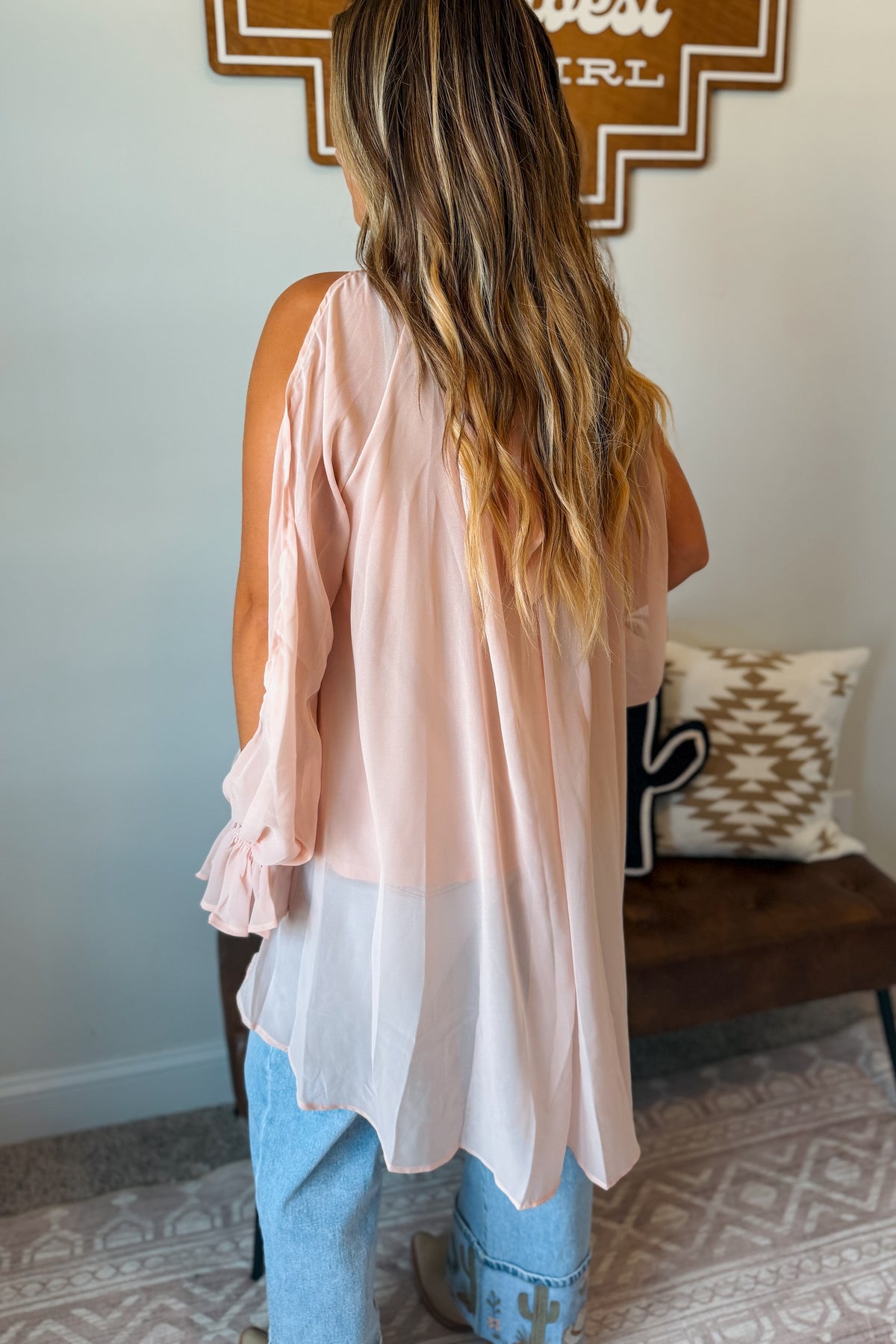 A woman shown from behind wearing a light pink sleeveless top with a sheer overlay and ruffled cuffs, paired with light blue jeans in an indoor setting.