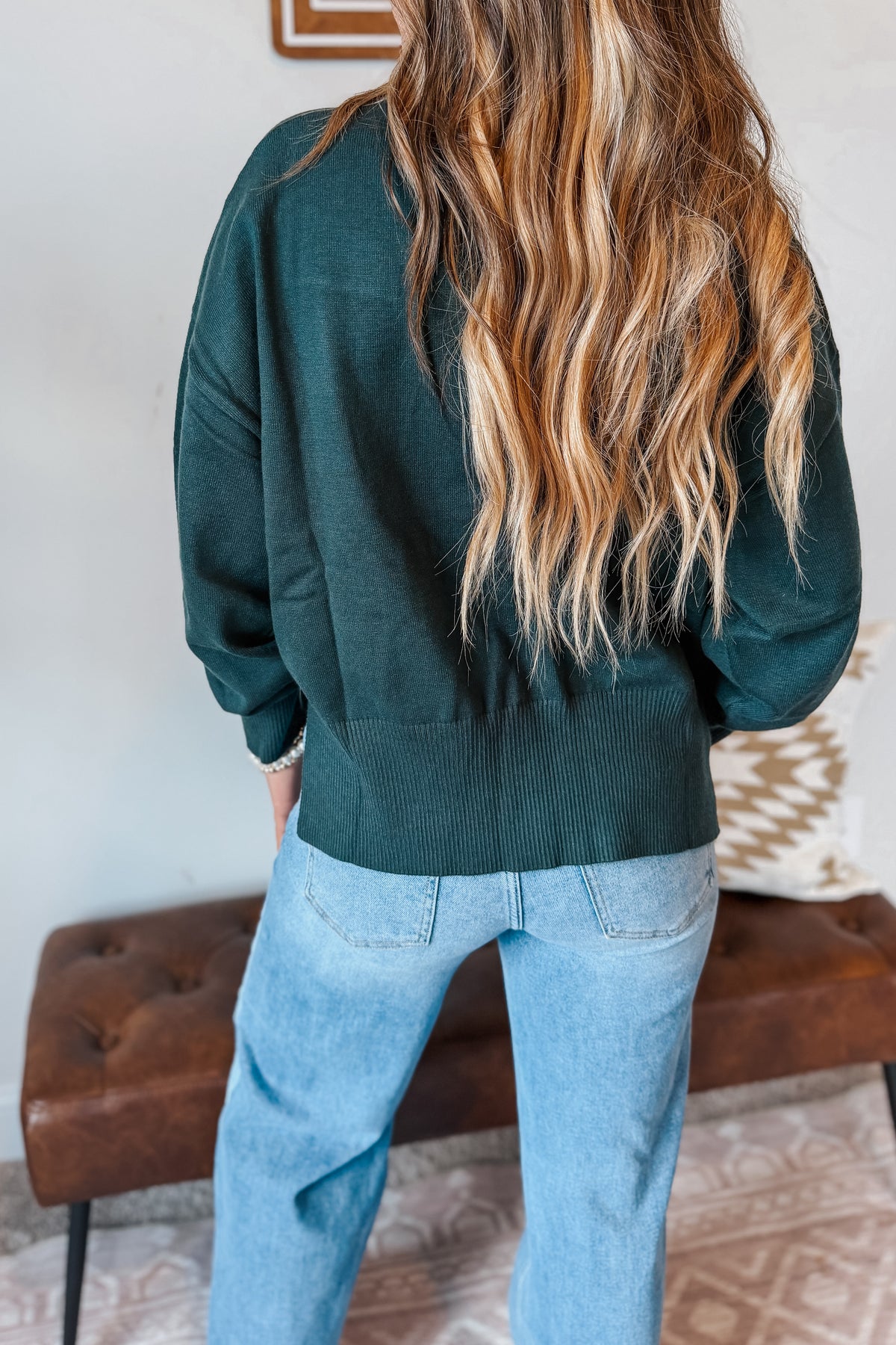 A woman viewed from behind wearing a dark forest green knit sweater and light blue jeans, standing in front of a brown leather bench in an indoor setting.