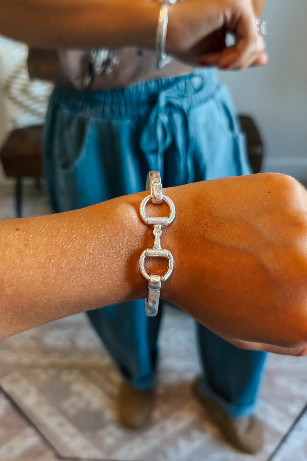 A close-up of a person's wrist wearing a silver horse-bit style link bracelet, with their teal drawstring sweatpants and bare arms visible in a softly blurred indoor setting.