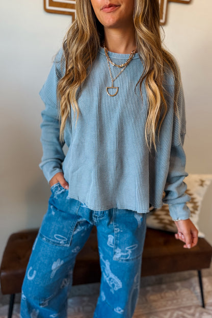 A woman wearing a light blue waffle knit long-sleeve top and blue distressed jeans, standing indoors.