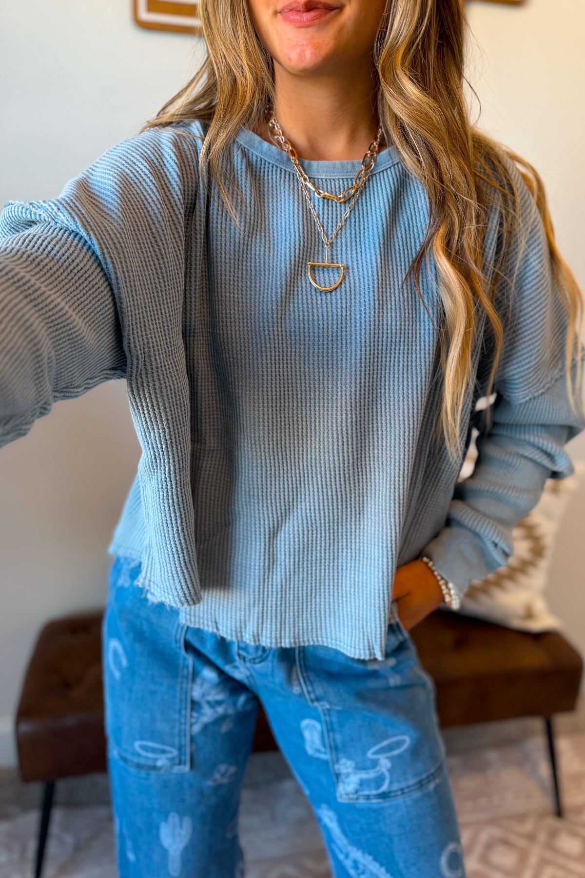 A woman wearing a light blue waffle knit long-sleeve top and blue jeans with white cactus prints, accessorized with layered gold necklaces. 