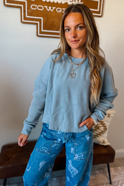 A woman wearing a light blue ribbed long-sleeve crop top and blue distressed jeans, standing in a home interior setting.