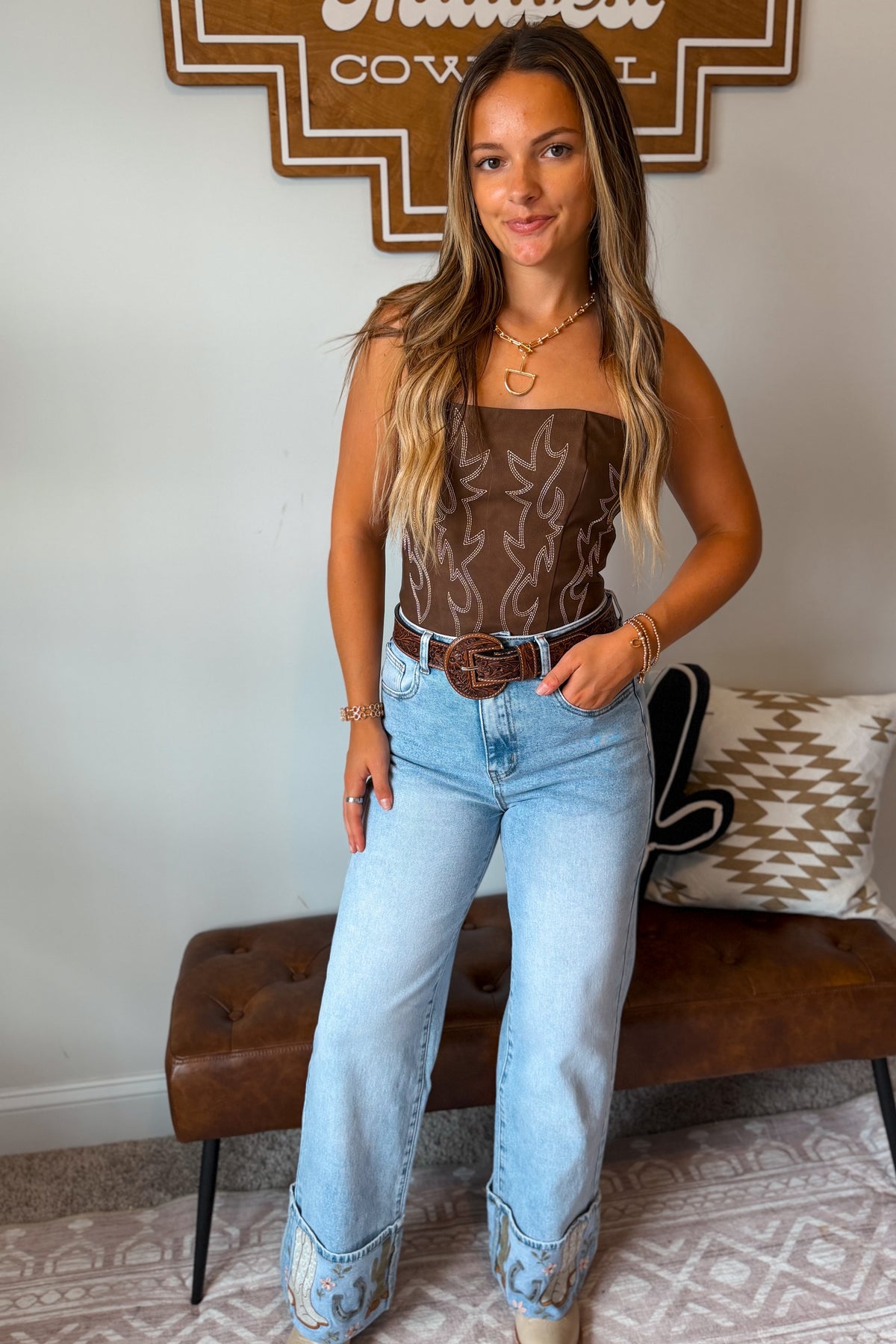A woman modeling a brown strapless western-style top with a boot stitch pattern, paired with light-wash blue jeans, a brown belt, and western boots,