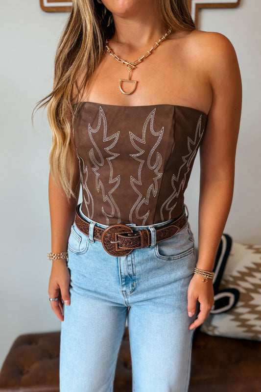 A woman wearing a brown strapless tube top with white western-style embroidery, paired with light blue jeans and a brown belt.