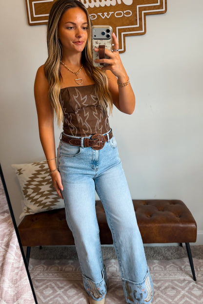 A woman takes a mirror selfie wearing a brown strapless top with a western boot stitch pattern, a brown leather belt, and light-wash blue jeans