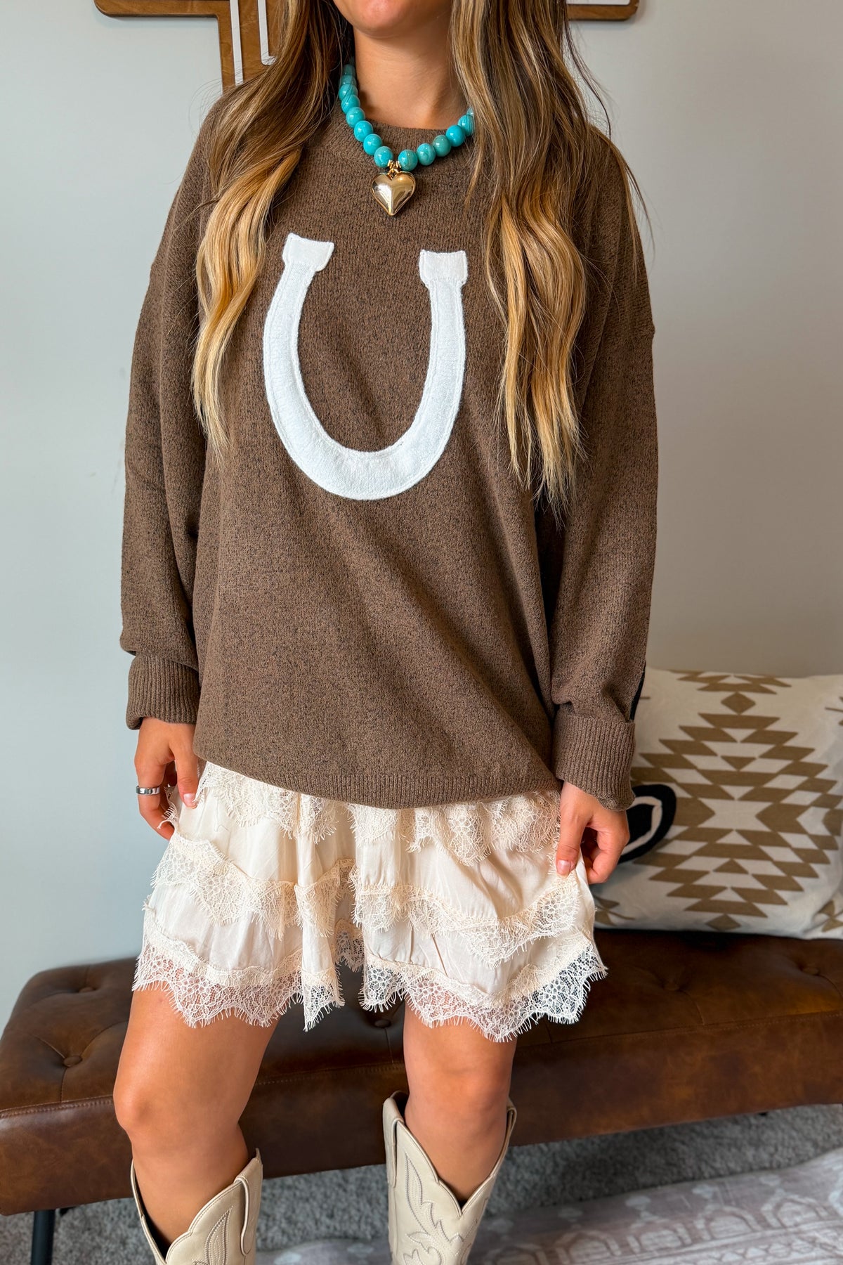 A woman wearing a brown knit sweater with a large white horseshoe graphic on the front, layered over a cream lace skirt and paired with white cowboy boots. She is wearing a turquoise necklace in an indoor setting.