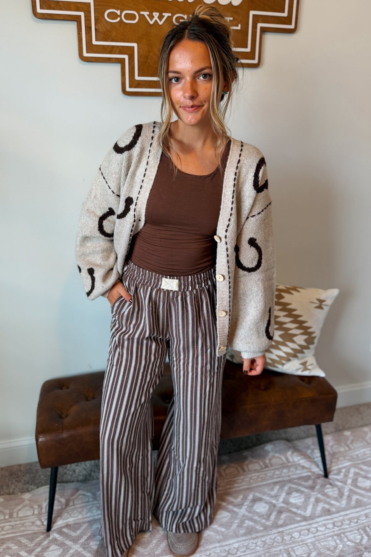 A woman stands indoors wearing a cream knit cardigan with black abstract motifs over a brown ribbed tank top and brown-and-beige vertically striped wide-leg pants. She is posed in front of a brown tufted bench with a patterned pillow, against a light wall with a decorative sign above.