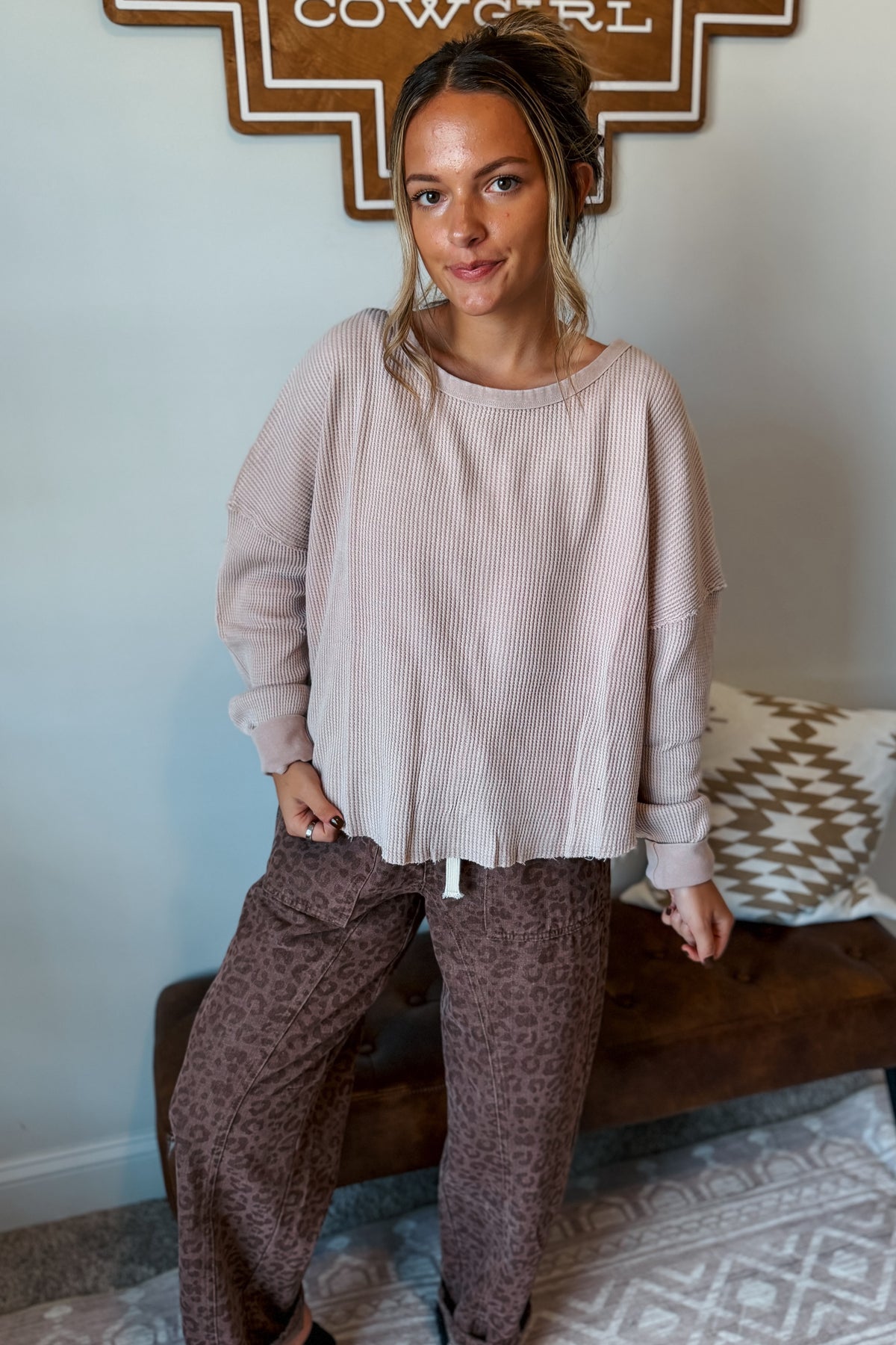A woman wearing a light pink ribbed long-sleeve top and brown leopard-print pants, sitting on a wooden bench in a home setting.